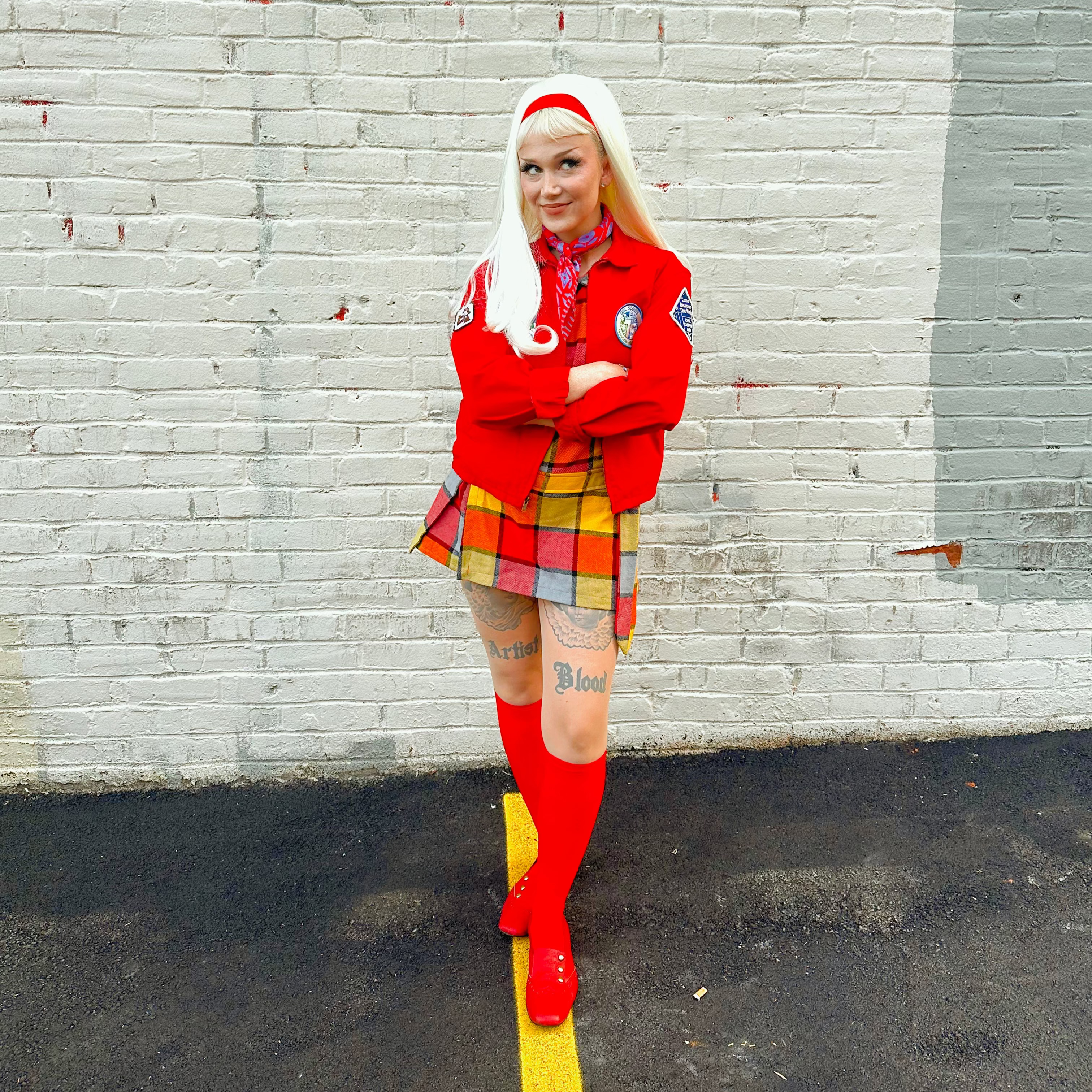 Woman with long blonde hair in a red outfit with plaid skirt standing against a light gray brick wall.